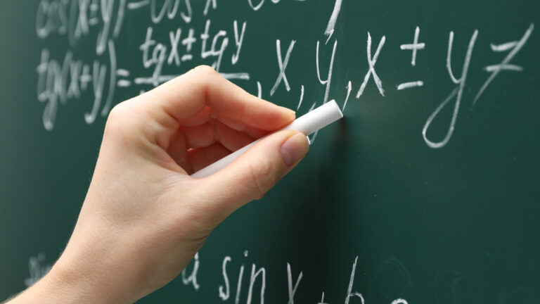 A hand with chalk calculating math problems on a classic green chalkboard.